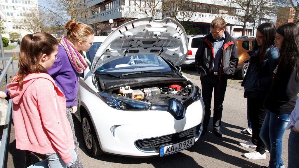 Schülerinnen betrachten mit Professor offene Motorhaube eines E-Fahrzeugs